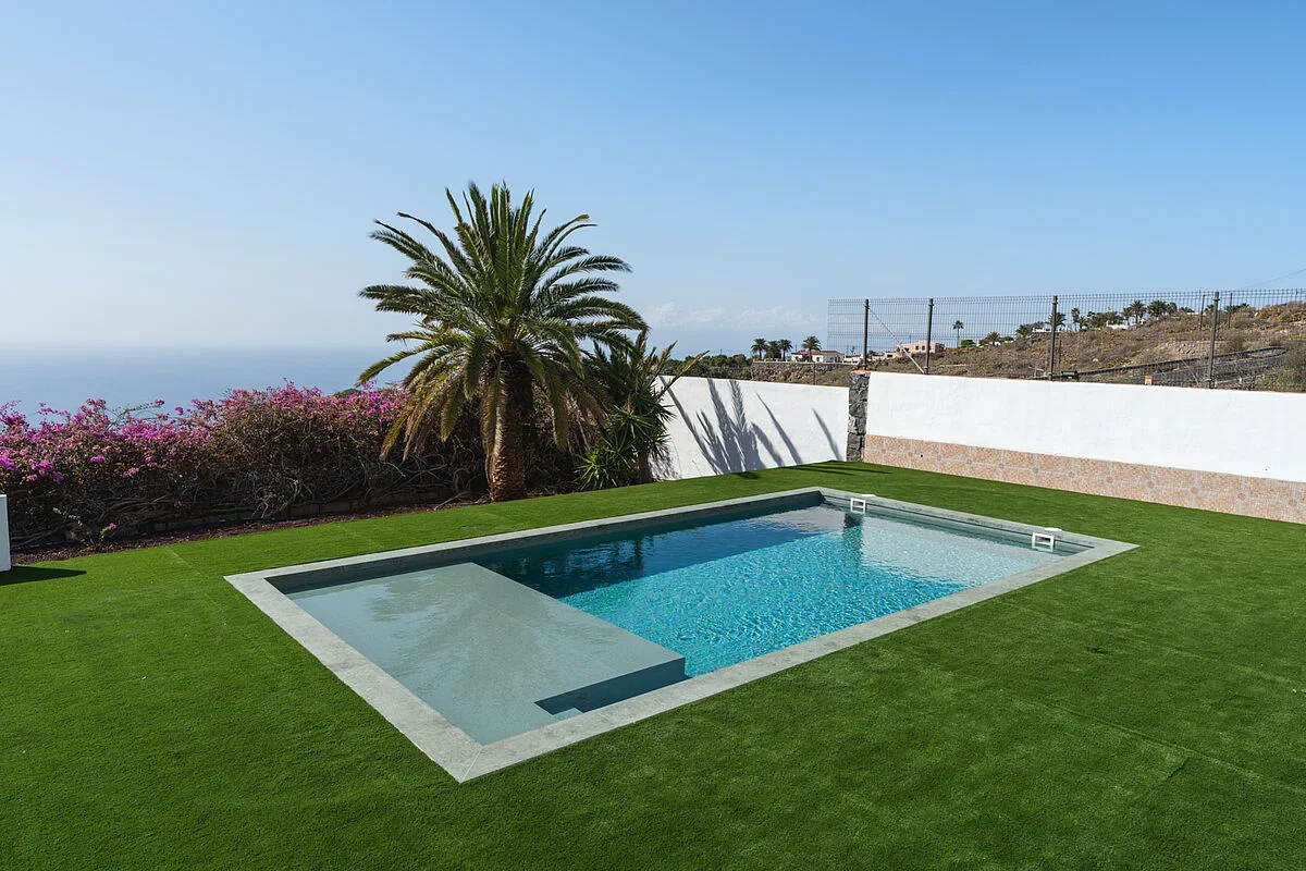 Piscine avec membrane armée RENOLIT ALKORPLAN Vogue Tropical, eau turquoise sur pelouse synthétique face à la mer et aux palmiers